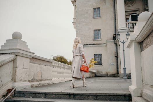 A woman wearing a white coat and carrying a red purse walks down a stone staircase. The scene is set in a city, with a large building in the background. The woman is in a hurry