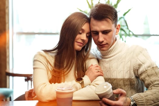 A couple sits close together at a wooden table in a brightly lit cafe, exchanging smiles over steaming cups of perhaps coffee or tea. They appear relaxed and engaged in a cheerful conversation, with natural light filtering through the space creating a warm and inviting atmosphere.