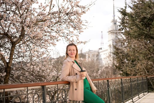 A woman is standing on a ledge with a cup of coffee in her hand. She is wearing sunglasses and a tan coat. The scene is set in a park with cherry blossoms in bloom. The woman is enjoying the view