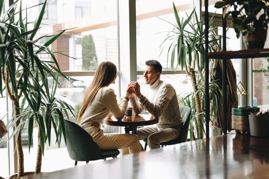 A man and a woman are seated across from each other in a sunlit cafe, holding hands and gazing into each others eyes, creating a moment of intimacy. The setting exudes warmth and tranquility, with indoor plants contributing to the serene atmosphere.