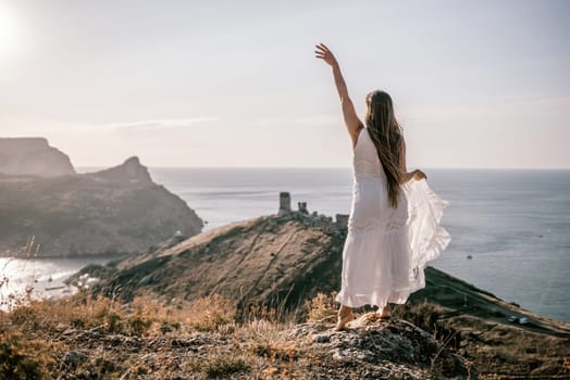 A woman in a white dress stands on a rocky hill overlooking the ocean. She is smiling and she is happy