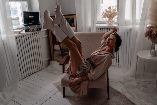 A woman is sitting in a chair with her legs spread apart. She is wearing white boots and a white dress. The room is decorated with a white color theme
