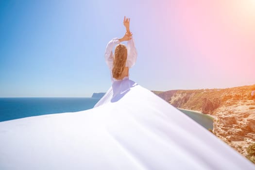 woman sea white dress. Blonde with long hair on a sunny seashore in a white flowing dress, rear view, silk fabric waving in the wind. Against the backdrop of the blue sky and mountains on the seashore