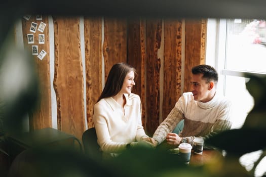 A woman and a man are sitting at a wooden table in a warmly lit cafe, their faces animated in what appears to be a relaxed conversation. The woman holds a paper cup, possibly containing coffee, as they both enjoy a comfortable moment together, surrounded by an ambiance of casual chatter.