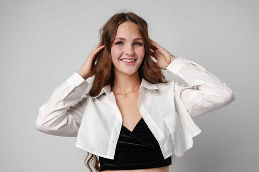 A young woman stands with a beaming smile, touching her hair lightly as she poses casually. She wears a stylish white jacket over a black top, complemented by a simple necklace, against a neutral grey backdrop.