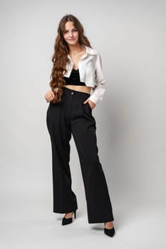 A young woman with flowing, wavy brown hair showcases her casual fashion sense, wearing an unbuttoned white shirt over a black crop top, paired with a black bottom. Her pose exudes confidence as she gazes off-camera, standing elegantly against a plain gray background.