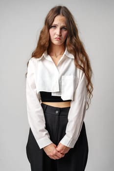 A young woman with flowing long brown hair stands with her hands in her pockets, donning a white shirt and black trousers. She gazes into the camera with a mixed expression of contemplation and seriousness, set against a plain gray backdrop.