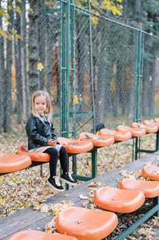 Little girl sits on a seat on a playground surrounded by a mesh fence. High quality photo