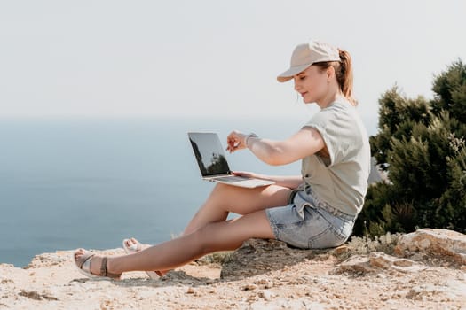 Digital nomad, woman in the hat, a business woman with a laptop sits on the rocks by the sea during sunset, makes a business transaction online from a distance. Freelance, remote work on vacation.