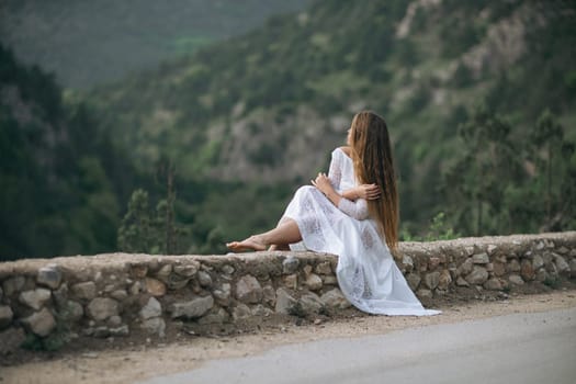 A woman in a white dress is sitting on a stone wall overlooking a mountain. The scene is serene and peaceful, with the woman's long hair blowing in the wind. Concept of calm and tranquility
