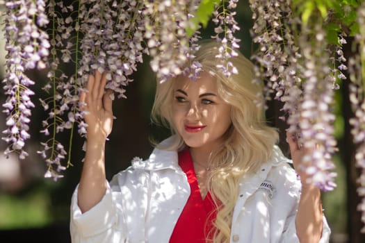 A blonde woman is standing in front of purple flowers. She is wearing a white jacket and red shirt