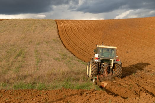 A tractor actively plowing a curved field with a stormy sky overhead, showcasing the dramatic contrast of agriculture and nature.