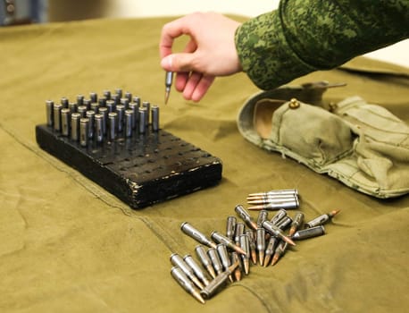 A soldiers hand is carefully reaching into a wooden box to hold a single bullet, while scattered bullets are arranged on an olive drab cloth surface, creating a compelling sense of depth and focus.