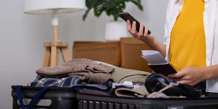 Young traveler asian woman using smartphone to book a hotel while preparing travel suitcase before going on summer vacation. Online booking concept.