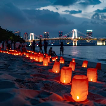 Illuminated paper lanterns line a sandy beach at twilight, with a beautifully lit suspension bridge and city skyline in the background. Japanese Marine Day Umi no Hi also known as Ocean or Sea Days.