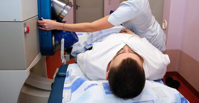 A young male patient is lying on a table getting a medical procedure done. A nurse is assisting him. The patient is wearing a white shirt and the nurse is wearing a white lab coat.