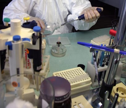 The scientist is meticulously working in a biosafety cabinet, wearing protective mask, gloves, and lab coat. Using a Bunsen burner, test tubes, and various lab equipment for experiments.