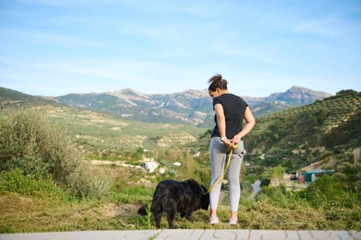 Full length portrait of a woman in sportswear, walking her dog on leash in the nature. Multi ethnic enjoying walk with her pedigree purebred black cocker spaniel dog in the mountains nature outdoors