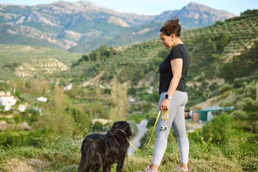 Full size portrait of a woman in sportswear, walking her dog on leash in the nature. Multi ethnic enjoying walk with her pedigree purebred black cocker spaniel dog in the mountains nature outdoors