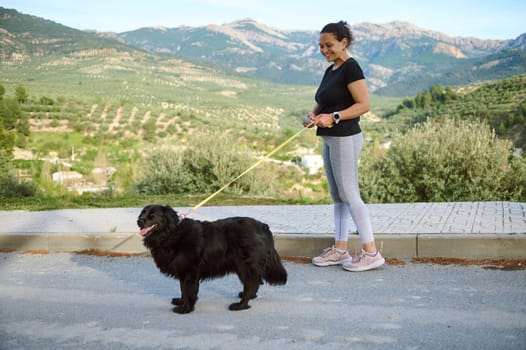 View from the side of a woman in sportswear, walking her dog on leash in the nature. Multi ethnic enjoying walk with her pedigree purebred black cocker spaniel dog in the mountains nature outdoors