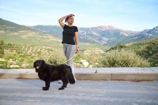 Beautiful active young woman in sportswear, looking way, walking her dog on leash in the nature, enjoying walk with her pedigree purebred black cocker spaniel dog in the mountains nature outdoors