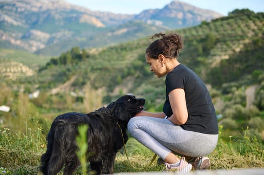 Affectionate young woman embracing her pedigree black cocker spaniel pet dog while walking him in the mountains nature. Playing pets and people concept