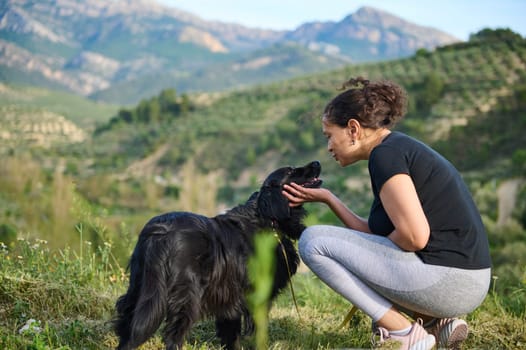 Affectionate young woman talking with her companion dog, embracing her pedigree black cocker spaniel pet while walking him in the mountains nature outdoors. Playing pets and people concept