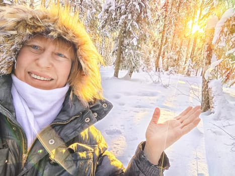 A cheerful middle aged woman in a winter coat with fur, scarf taking selfie outdoors on nature in a forest or park in beautiful sunny day