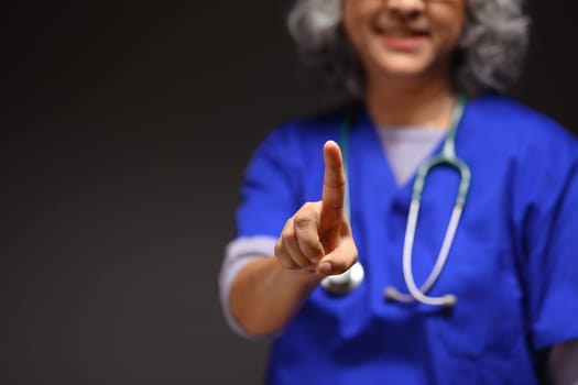 Doctor with stethoscope standing against black background and pointing towards the camera.