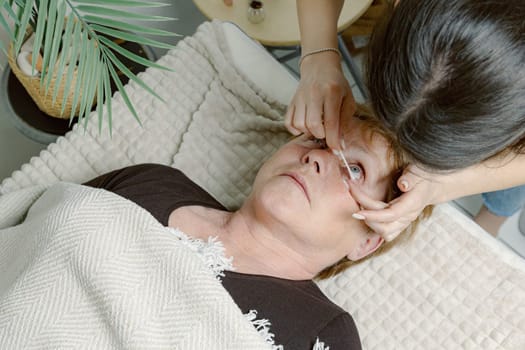 One Caucasian young brunette cosmetologist wipes the eyelashes of the lower eyelid with a cotton swab to an elderly female client, preparing them for the lamination procedure, which is lying on the cosmetology table in a home beauty salon, side view close-up.