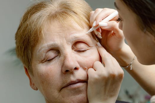 One young Caucasian girl cosmetologist draws a contour with a white marker on the left eyebrow of an elderly beautiful woman with her eyes closed, sitting in a home beauty salon, close-up side view.