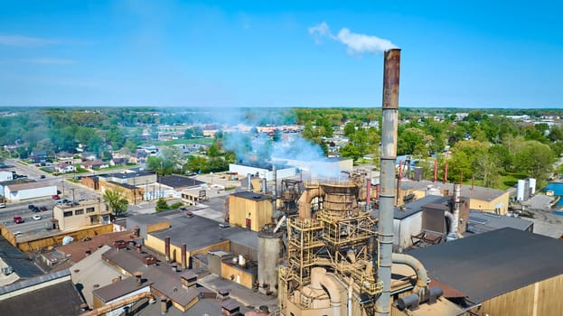 Aerial view of an active industrial complex in Warsaw, Indiana, with smokestack emissions and surrounding suburban homes.