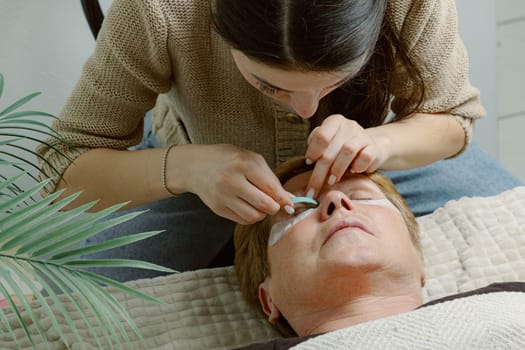 One Caucasian young beautiful brunette girl cosmetologist sticking a blue silicone eyelash stand for the right eye of an elderly client who is lying on a cosmetology bed in a home beauty salon, top side close-up view.
