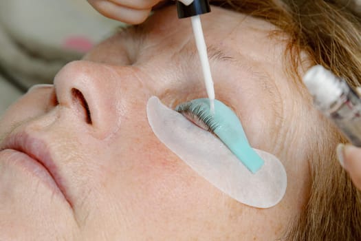 One Caucasian young unrecognizable girl cosmetologist applies transparent glue to the eyelashes of the left eye with a brush, pulling them onto a silicone stand of an elderly female client who is lying on a cosmetology bed in a home beauty salon, top side view very close-up.