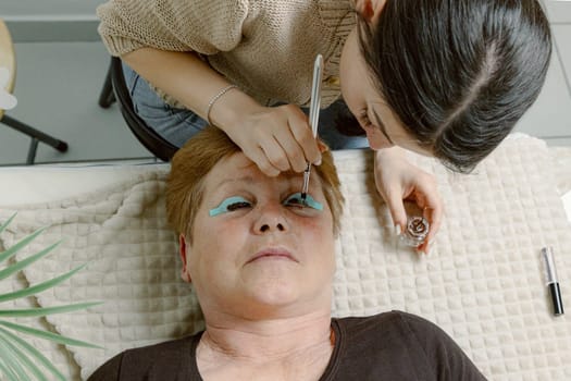 One Caucasian young beautiful brunette cosmetologist applies brown paint with a brush to the eyelashes of the left eye of an elderly woman client who is lying on the cosmetology table in a home beauty salon, flat lay close-up.