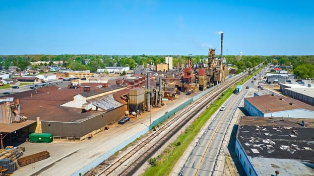 Aerial view of a bustling industrial zone in Warsaw, Indiana, with smokestacks and railroads under a clear blue sky.