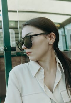 Portrait of one beautiful young Caucasian brunette girl in sunglasses and stylish clothes looking at nature while sitting in an open carriage of a tourist train on a sunny spring day in a public park, close-up side view.