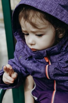 Portrait of one beautiful little Caucasian brunette baby girl in a lilac jacket with a hood, sitting in an open carriage of a tourist train, looking to the side and crossing her fingers, driving through a nature reserve on a sunny spring day in a public park, close-up side view.