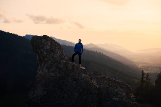 A man is perched on a rock in the highlands, surrounded by mountain ranges as the sun sets, creating a breathtaking atmospheric phenomenon in the sky