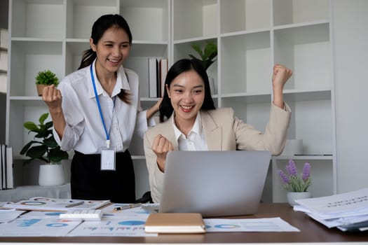 Two Asian businesswomen celebrating success while working on a laptop in the office. Concept of achievement and teamwork.