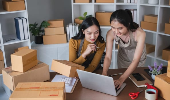 Two women working on laptop and packing boxes in warehouse.