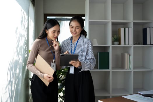 Two Asian businesswomen discussing work using a tablet in a modern office. Concept of teamwork and professional collaboration.