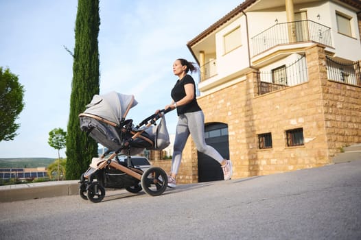 Happy woman performing morning jog, pushing a baby pram running on the city street and enjoying her active lifestyle during maternity leave. People. Motherhood and babyhood concept