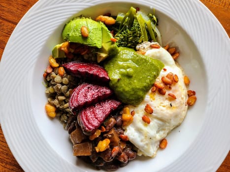 Fresh, nutritious rice bowl with sunny side up egg, avocado slices, pesto, lentils, beans, broccoli, and tempeh pieces on a rustic wooden table.