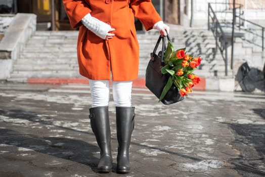 A beautiful young woman in a orange coat and rubber boots walking down the street with a bouquet of tulips in spring city