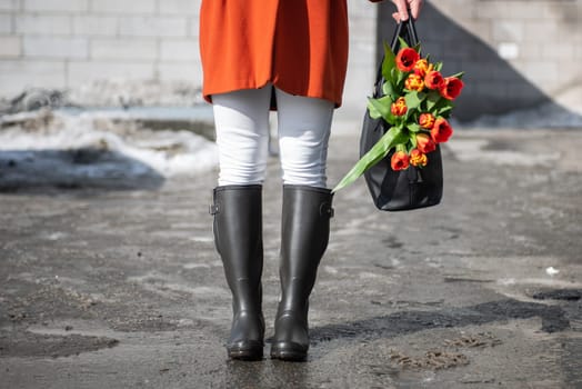 A beautiful young woman in a orange coat and rubber boots walking down the street with a bouquet of tulips in spring city