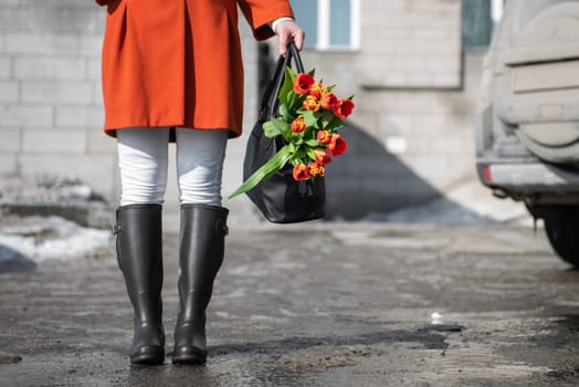 A beautiful young woman in a orange coat and rubber boots walking down the street with a bouquet of tulips in spring city