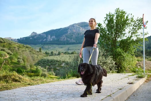 Multi ethnic happy smiling woman in sportswear, walking her dog on leash in the nature. Playing pets, people and animals concept