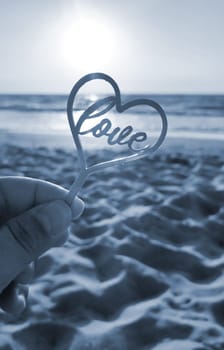 Person holding in fingers hand stick in shape heart and word Love on background sea, waves, blue sky in seashore on sunny summer day close-up. Concept love romance amour St Valentines Day. Blue color