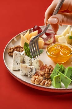 Woman piercing piece of blue cheese with fork, savoring delightful cheese platter traditionally served with nuts, honey, and grapes, presented on elegant plate against red background, cropped shot